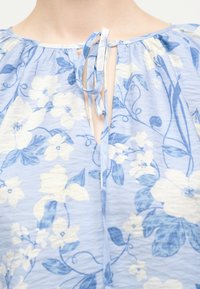 Λαιμόκοψη ανοιχτό μπλε μπλούζας με λευκά λουλούδια, κλείσιμο με δεσίματα και ρυμουλκούμενο ύφασμα γύρω από τον λαιμό σε ανοιχτόχρωμο τόνο δέρματος.