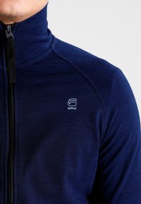 Giacca zip-up navy con colletto alto, realizzata in tessuto testurizzato. Presenta un piccolo logo bianco sul lato sinistro del petto. Zip nera.