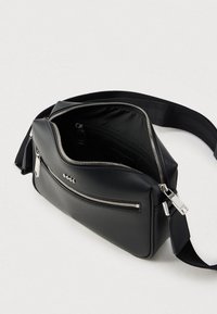 Μαύρη τσάντα χιαστί από υλικό με υφή, με ασημένια φερμουάρ, μπροστινή τσέπη και ρυθμιζόμενο λουρί. Στο εσωτερικό υπάρχει επιπλέον τσέπη με φερμουάρ.