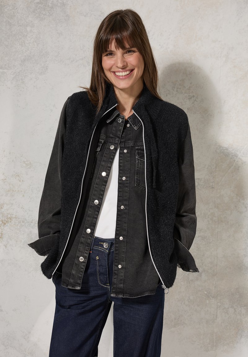 Giacca di jeans nera con colletto a pelo, texturizzato. Caratterizzata da hardware argentato, profilature bianche e un design a strati su una maglietta bianca.