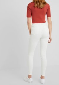 Kvinna som bär figursydda vita jeans, vita sneakers och en kortärmad topp i rostfärgad nyans, stående mot en enkel ljus bakgrund.