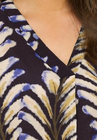 Blusa con stampa floreale in una profonda tonalità di viola. Presenta un scollo a V e un tessuto fluente con motivi blu e gialli che ricordano le foglie. Morbida al tatto.