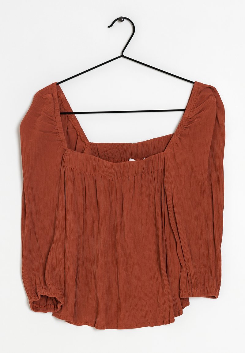 Blouse couleur rouille avec un large décolleté et des manches longues. En tissu texturé avec des détails froncés à la taille et aux poignets des manches.