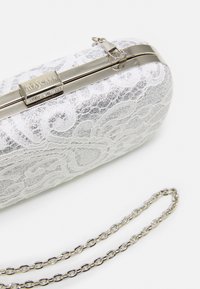 Pochette blanche scintillante avec overlay en dentelle et monture métallique argentée, fermoir gravé "MASCARA" et bandoulière en chaîne argentée sur surface blanche.