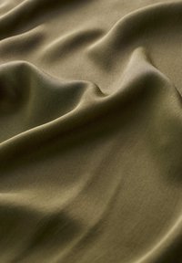 Olivově zelená látka s hladkým texture, drapovaná tak, aby ukazovala jemné vlny a subtilní lesk, zdůrazňující její fluidnost a elegantní záhyby.