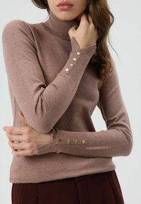 Bruine, aansluitende coltrui van zachte stof, met drie gouden knopen op elke manchet en geribbelde details bij de kraag en zoom.