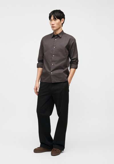 Νεαρός άνδρας στέκεται με τα χέρια στις τσέπες, φοράει σκούρο καφέ πουκάμισο, μαύρο παντελόνι με φαρδιά μπατζάκια και καφέ casual παπούτσια σε απλό φόντο.