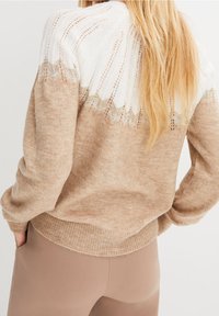 Beige Pullover mit einer weißen, gestrickten Schulterpartie im Spitzenmuster. Weiche Textur und gerippter Saum, kombiniert mit eng anliegenden beigen Hosen.