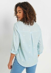 Light blue shirt made of textured fabric, featuring white and yellow daisy embroidery, long sleeves rolled up, and a back pleat detail.