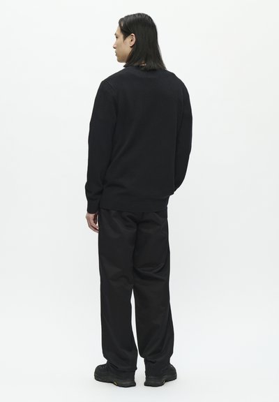 Suéter de punto negro con escote redondo, combinado con pantalones negros holgados y zapatillas negras gruesas, visto desde la parte de atrás sobre un fondo blanco.