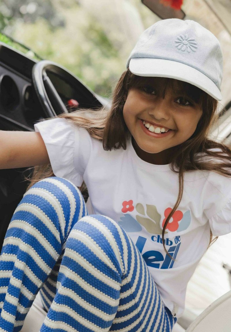 Kind draagt een wit T-shirt met een kleurrijk bloemenpatroon en blauw-wit gestreepte legging, gecombineerd met een lichtblauwe pet.