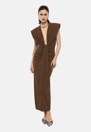 Donna che indossa un lungo vestito marrone senza maniche con scollo profondo a V e davanti arricciato, abbinato a scarpe a punta con righe zebrate e gioielli dorati.