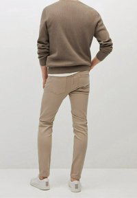 Beżowy prążkowany sweter z długimi rękawami, zestawiony z beżowymi spodniami o wąskim kroju i białymi trampkami. Widok z tyłu, na neutralnym tle.