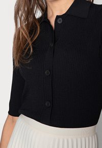 Svart ribbad kortärmad topp med krage, som har sex svarta knappar framtill och ett kontrasterande lätt plisserat midjeband på kjolen.