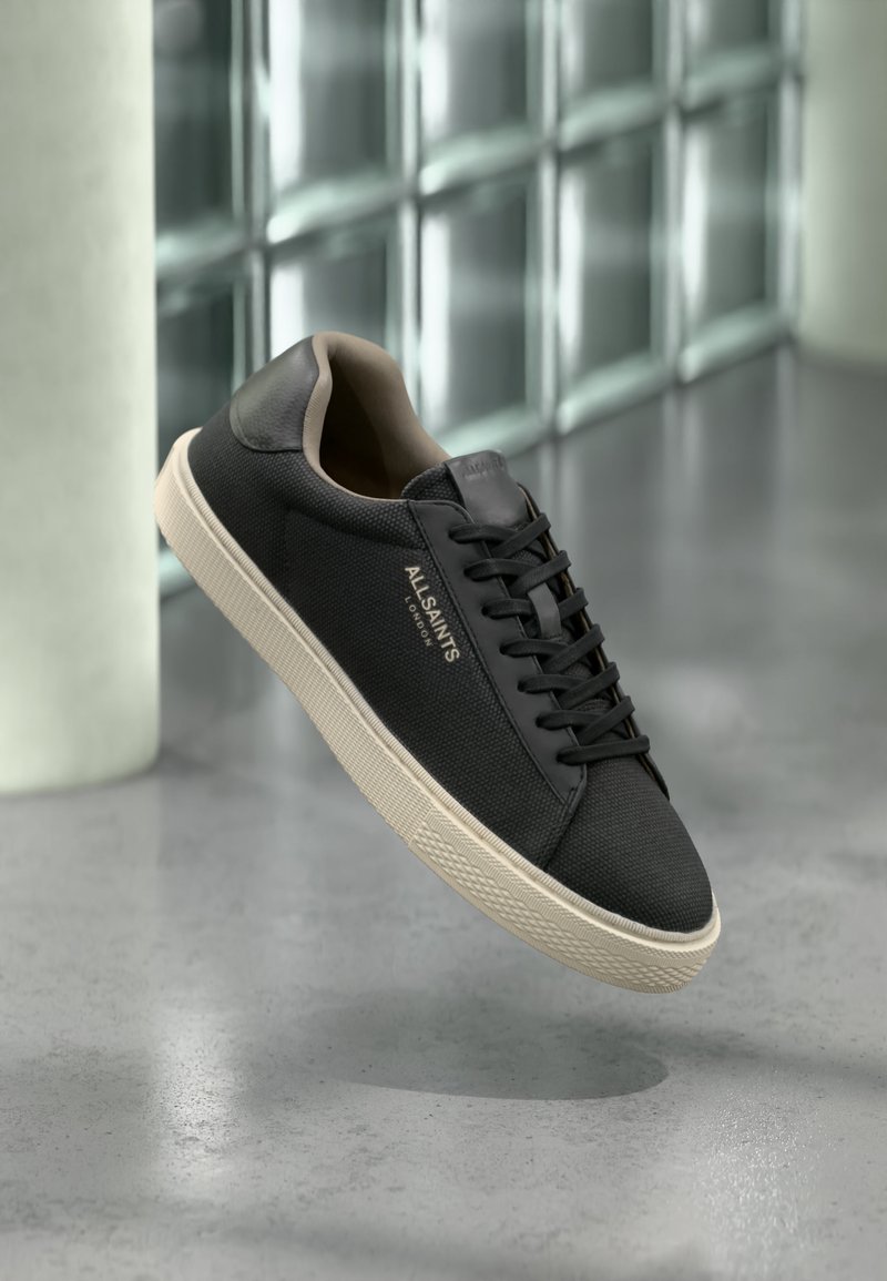 Μαύρο sneaker χαμηλού τύπου με μπεζ σόλα και κορδόνια, λογότυπο "ALLSAINTS LONDON" στο πλάι, αιωρείται πάνω από γυαλισμένο δάπεδο από σκυρόδεμα με φόντο τοίχο από γυάλινους κύβους.