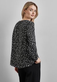 Blusa a maniche lunghe bianco e nero con motivo geometrico astratto, tessuto morbido, scollatura rotonda e dettaglio a pieghe sul retro.