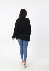 Maglione nero oversize con maniche lunghe, indossato sopra jeans skinny azzurri. Tacchi trasparenti visibili. Sfondo semplice.