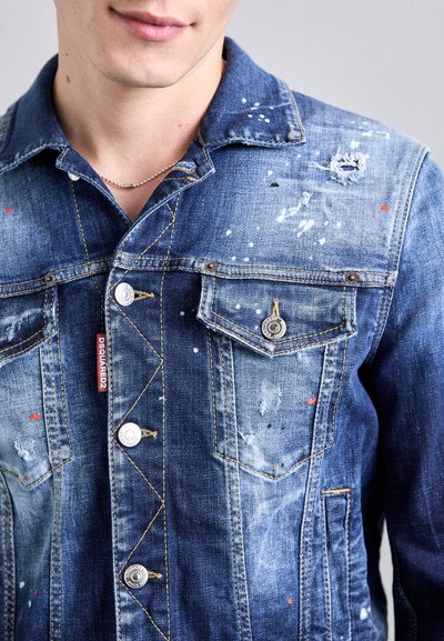 Chaqueta de denim en azul con detalles desgastados y salpicaduras de pintura. Presenta botones frontales y bolsillos con costuras visibles y una etiqueta de marca.