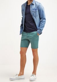 Denim overhemd met lange mouwen over een marineblauw t-shirt, gecombineerd met groene shorts en witte boot schoenen. Korte mouwen, lichte stof, casual pasvorm.