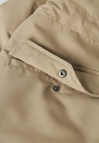 Nabij een beige stof met een knoopsluiting en een flapzak, zichtbaar stikwerk, met een zachte, gladde textuur en gedempte kleur.