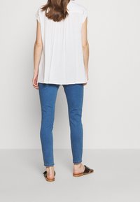 Top blanco de manga corta con pliegues en la espalda, combinado con jeans ajustados azules y sandalias negras sobre un fondo neutro.