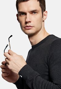 Czarny dzianinowy sweter z okrągłym dekoltem, z delikatnym logo. Osoba trzyma okulary w czarnej oprawie z metalowymi akcentami.