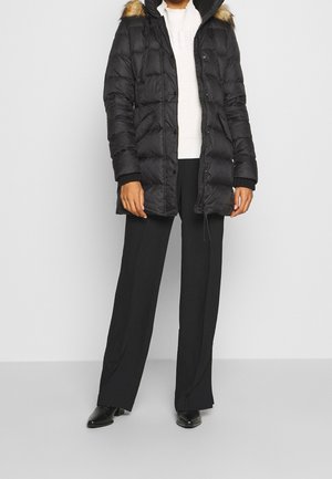 Personne portant un parka noir matelassé avec capuche en fourrure, un pull blanc, un pantalon noir à jambes larges et des chaussures pointues noires, debout sur un fond blanc.
