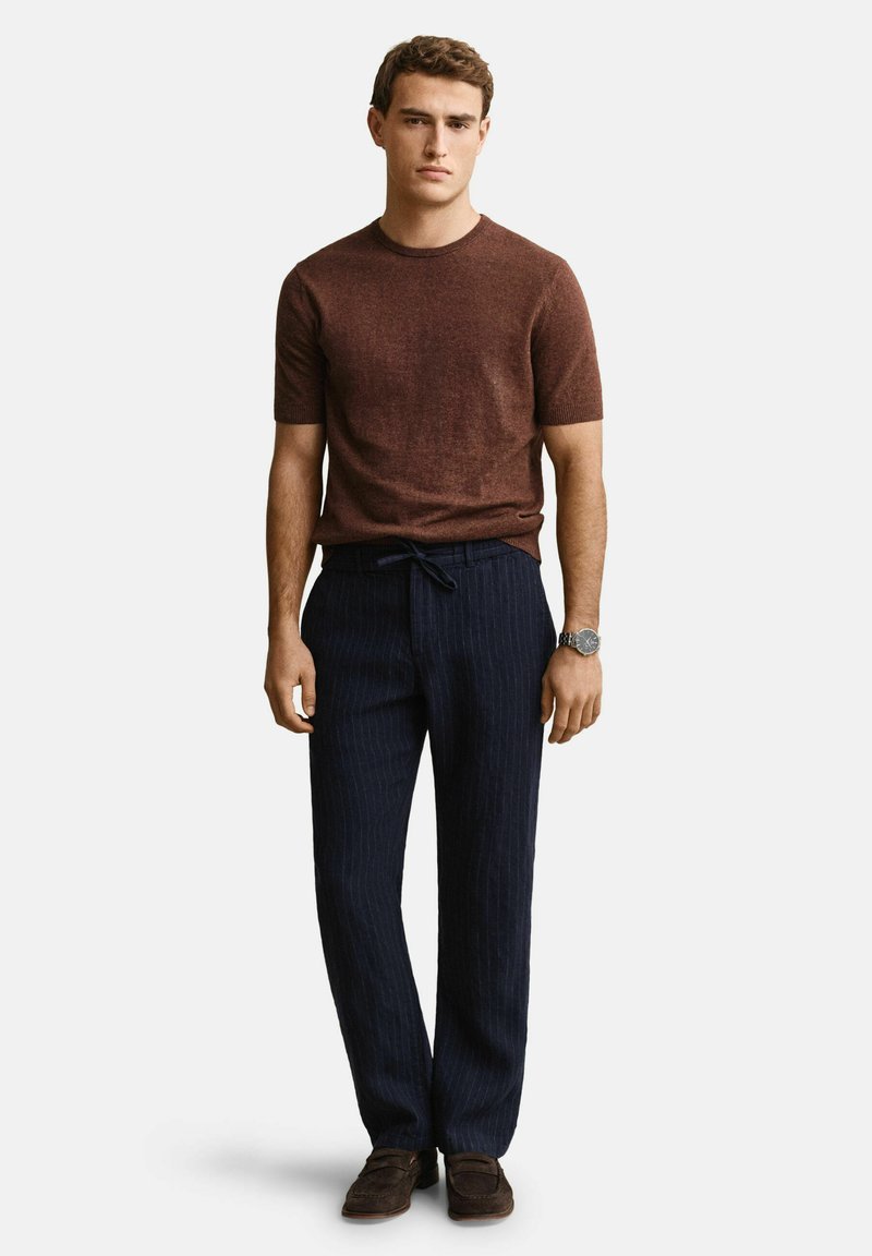 Junger Mann mit braunem Kurzarmpullover, marineblauen Nadelstreifenhosen mit Kordelzug, braunen Loafers und einer Armbanduhr, steht entspannt.