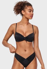 Bikini negro con un detalle de nudo en el frente en la parte superior y una parte inferior de corte bajo. Fabricado en tejido suave, con lados fruncidos en la parte inferior.