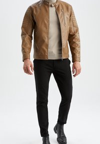 Uomo che indossa una giacca di pelle beige, un maglione beige, pantaloni neri slim e stivali Chelsea neri, in piedi contro uno sfondo bianco.