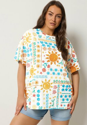 Femme portant un T-shirt oversize coloré avec des motifs de soleil, de lune et de fleurs, associé à un short en denim bleu clair.
