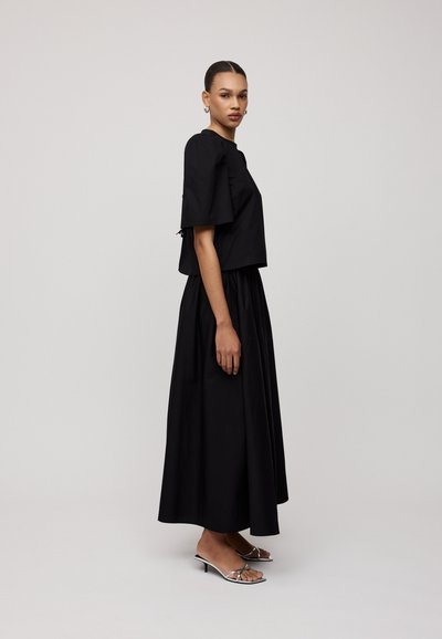 Zwarte, cropped top met korte mouwen gecombineerd met een vloeiende zwarte rok. Het model draagt metallic open-toe hakken, wat een slanke silhouet benadrukt.