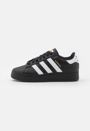 Czarny sneaker Adidas Superstar z białymi paskami, wzmocnionym noskiem, czarną podeszwą i złotym napisem "SUPERSTAR" po boku.