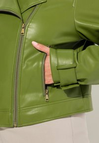 Veste en simili cuir vert avec fermeture éclair, col proéminent et poches latérales. Surface texturée et coupe ajustée avec des poignets accentués.