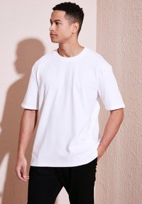 Tricou alb cu mânecă scurtă, realizat din bumbac texturat, cu decolteu rotund și manșete răsucite, purtat cu pantaloni negri, pe un fundal de perete deschis.