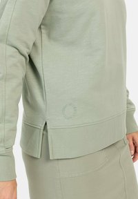 Lichtgroene sweatshirt met een ribbelzoom en zijsplitten, reliëfdetail "HOME SWEET EARTH" op de onderste linkerzijde, zachte textuur.
