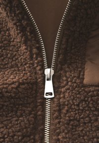 Veste en polaire marron avec une fermeture éclair en métal argenté, un extérieur texturé et une doublure lisse. Grande poche notable sur le côté droit.