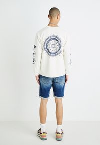Chemise crème à manches longues avec un design circulaire bleu marine au dos, associée à un short en denim roulé. Modèle portant des baskets colorées.