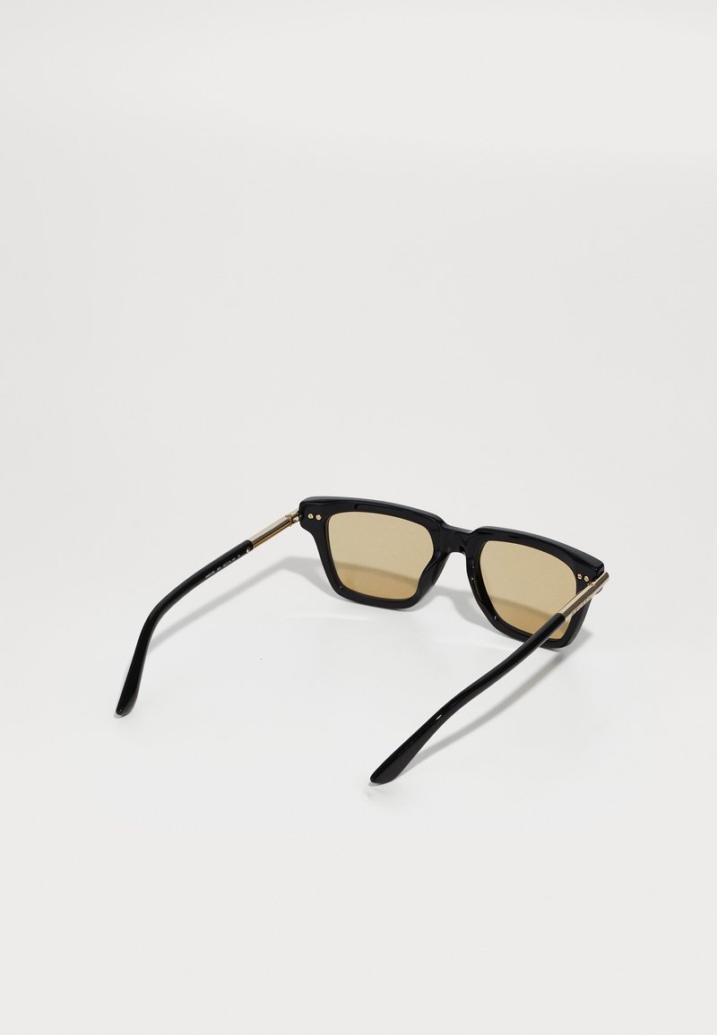 Schwarze Plastiksonnenbrille mit quadratischem Rahmen, braun getönten Gläsern und goldenen Metalldetails an den Bügeln. Glatte Oberfläche.