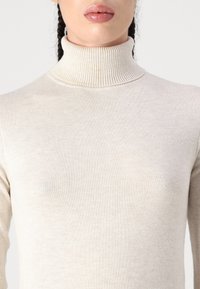 Ljust beige polokrage tröja med ribbad krage. Tillverkad av ett slätt stickat tyg med en figurnära silhuett. Subtil textur genom hela.