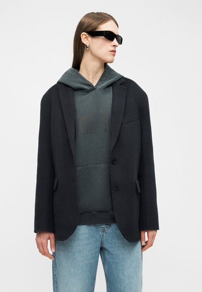 Ženska z ravnimi rjavimi lasmi, ki nosi črna sončna očala, srebrne obročke za ušesa, temen blazer čez temno kapuco in svetlo modre kavbojke.