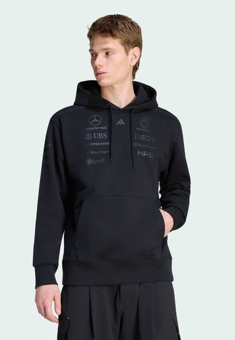 Zwarte hoodie gemaakt van zachte stof, met een voorzak in de stijl van een kangoeroezak en verschillende logo's in donkere tinten gedrukt op de voorkant en mouwen.