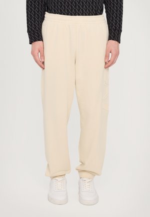 Beige sweatpants with a relaxed fit, elastic waistband, and side details in a matching matte texture, paired with white sneakers.