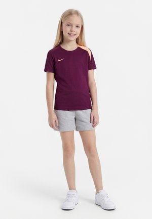 Jeune fille debout portant un t-shirt Nike violet avec un détail orange, un short gris et des baskets blanches, souriant à la caméra.