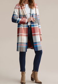 Vrouw in een lange geruite jas in rood, blauw en wit, over een witte top, donkere skinny jeans en tan suede enkellaarzen met blokhakken.