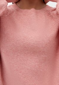Personne portant un pull en tricot rose avec des manches à motifs, ajustant ou tirant le col autour du cou.