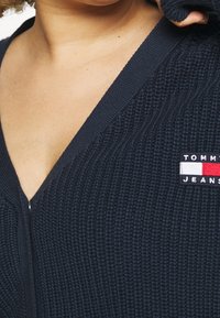 Granatowy sweter rozpinany z dzianiny, z dekoltem w serek i zamkiem błyskawicznym. Z przodu po lewej stronie małą naszywkę z logo w kolorach czerwonym, białym i niebieskim. Tkanina o fakturze.