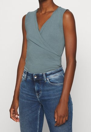 Bezrękawny, stonowany zielony top w stylu kopertowym połączony z niebieskimi jeansami o średnim stanie na stojącej osobie.