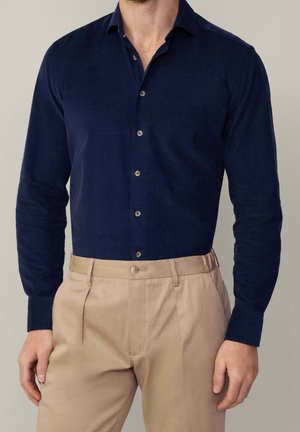Homme portant une chemise bleu marine cintrée à manches longues boutonnée, rentrée dans un pantalon beige plissé avec passants de ceinture.