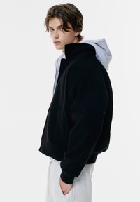 Schwarze Fleecejacke mit hohem Kragen, grauer Kapuzenpulli darunter, Kängurutasche und gerippte Bündchen, getragen über hellgrauen Hosen.